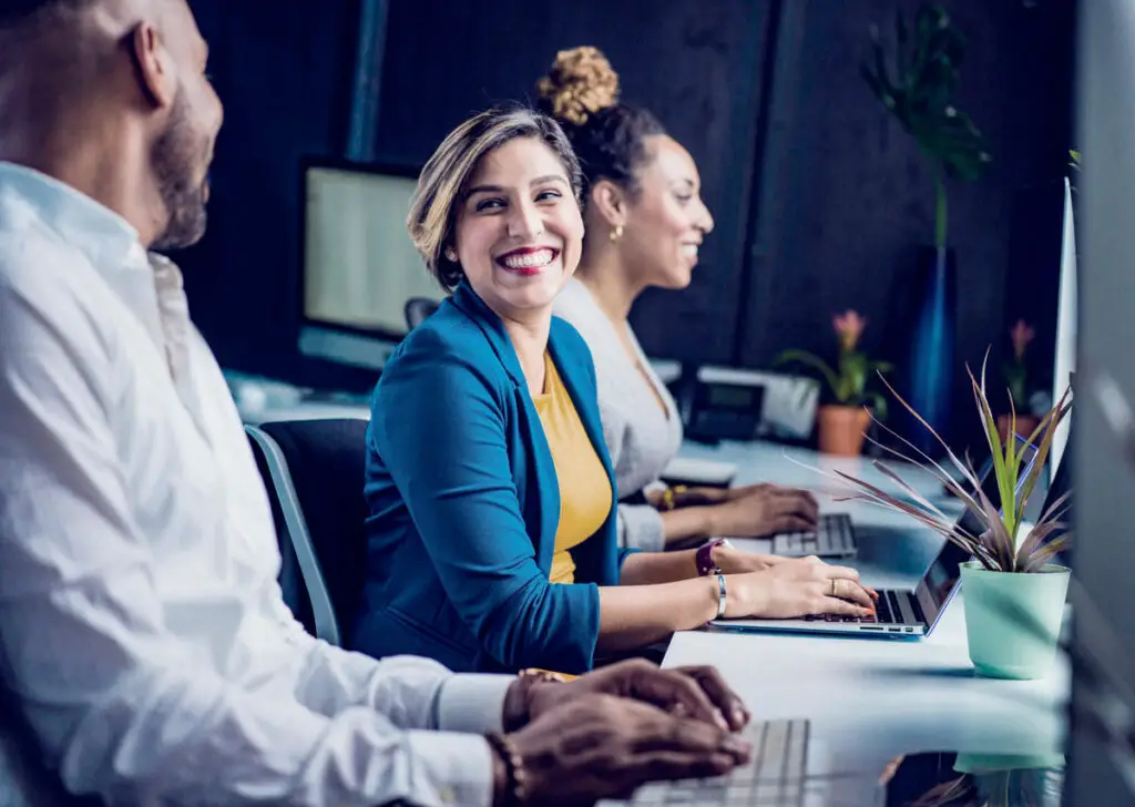 Three people sitting at a long desk working on laptops in a modern office with potted plants and large monitors