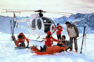 Mountain rescue team in red jackets assisting an injured skier on a stretcher in a snowy alpine area, with a white rescue helicopter parked nearby and ski equipment visible