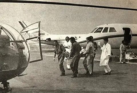 Group of people carrying a patient on a stretcher across an airport tarmac toward a helicopter, with a small aircraft parked in the background