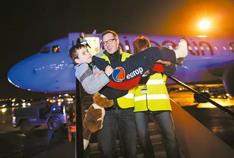 Two airport staff in high-visibility jackets assisting a child on an aircraft stairway at night, with an Air Berlin plane in the background and bright runway lights illuminating the scene