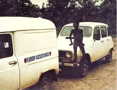 Two Europ Assistance service vehicles parked on a dirt road in a rural setting, with one person sitting on the hood of the second vehicle