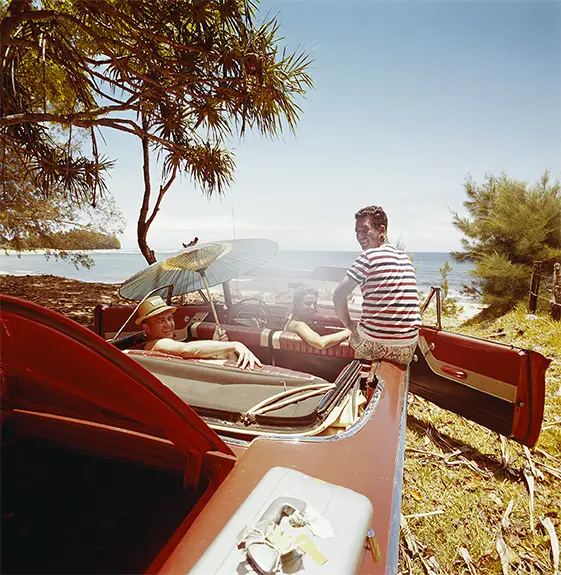 Open-top classic car parked under trees near a beach, with people relaxing inside and one person sitting on the car door, a parasol providing shade, and the ocean visible in the background