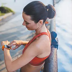 Person wearing a red sports top standing outdoors near a waterfront, checking a smartwatch while leaning on a railing