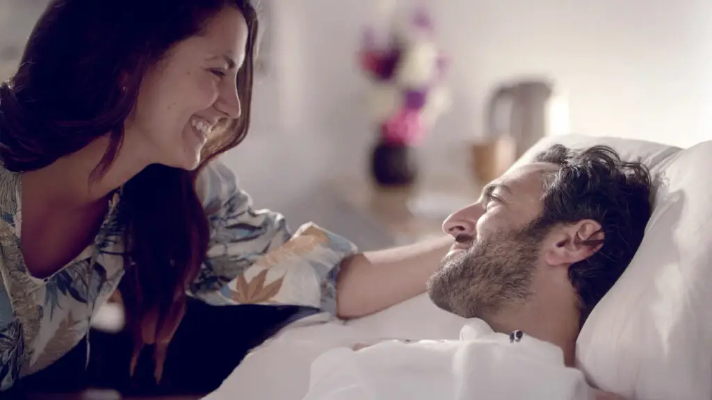 Person lying in a hospital bed covered with a white blanket, while another person leans close